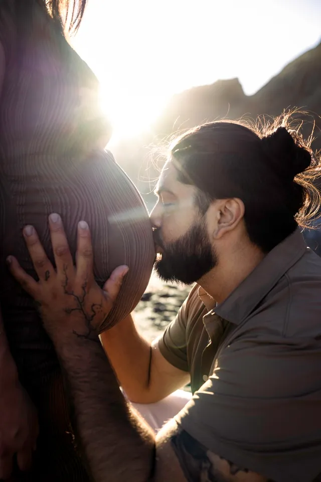 Partner kneeling to kiss a pregnant belly in golden backlight.