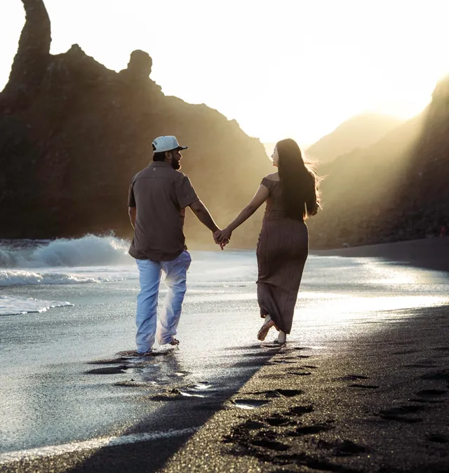 Couple holding hands on a black-sand beach at sunset.