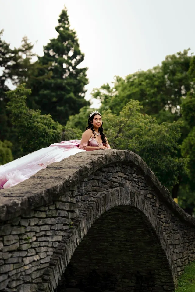 Quinceañera in a pink ball gown standing on a stone bridge, holding a bouquet.