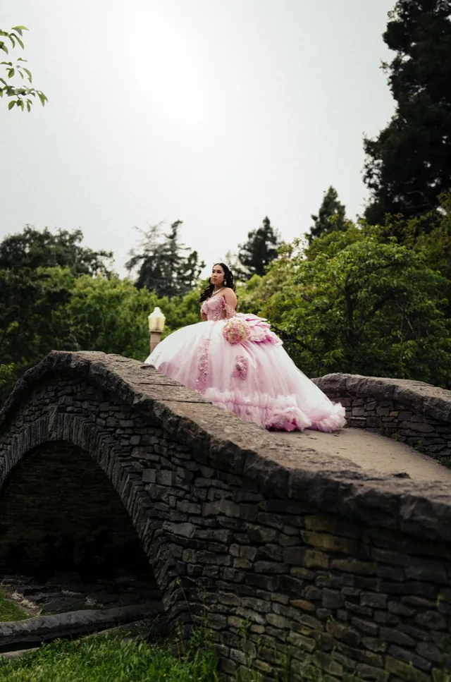 Quinceañera in a pink ball gown leaning on the wall of a stone bridge.